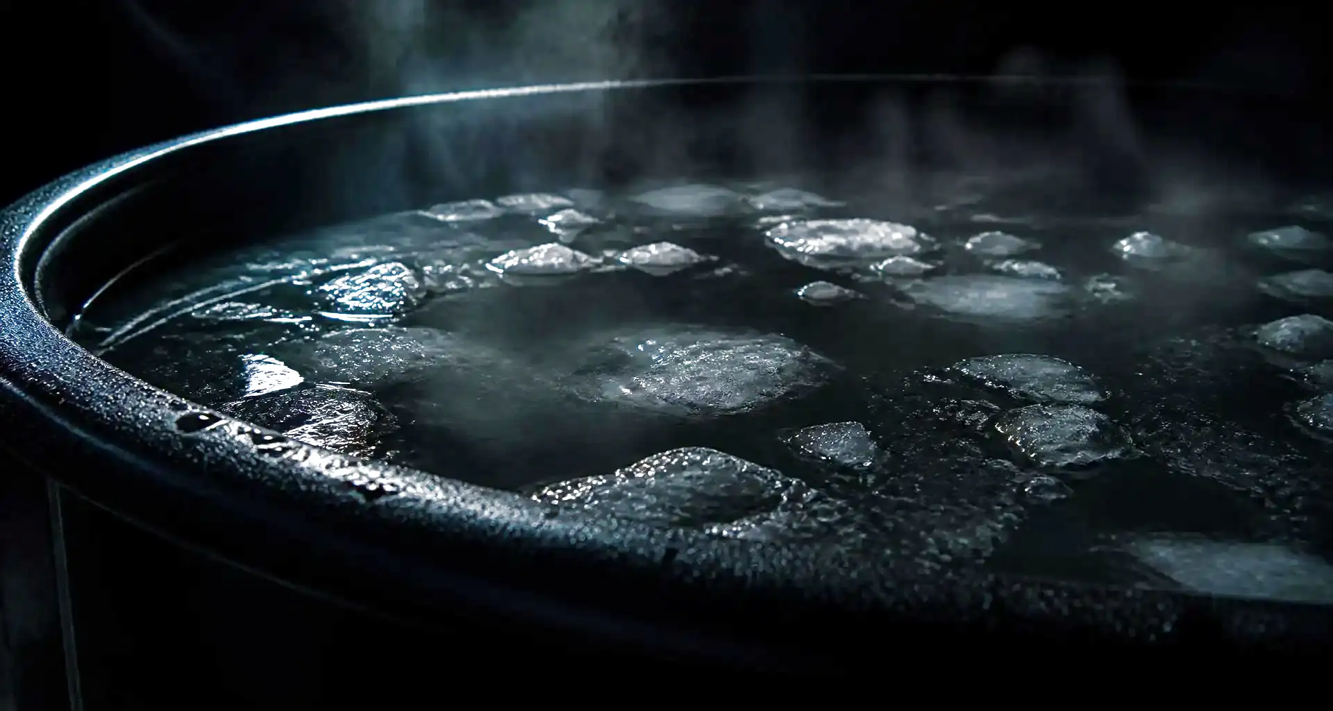 Ice floating on the black surface of a cold plunge tub, vapour rising